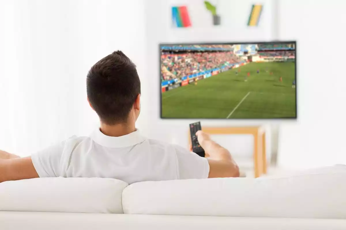 Un hombre sentado en un sofá de espaldas viendo fútbol en la televisión con un mando en la mano